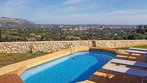 Ein Schwimmbecken mit Holzterrasse und Liegestühlen, umgeben von einer Steinmauer mit Blick auf eine Stadtlandschaft.