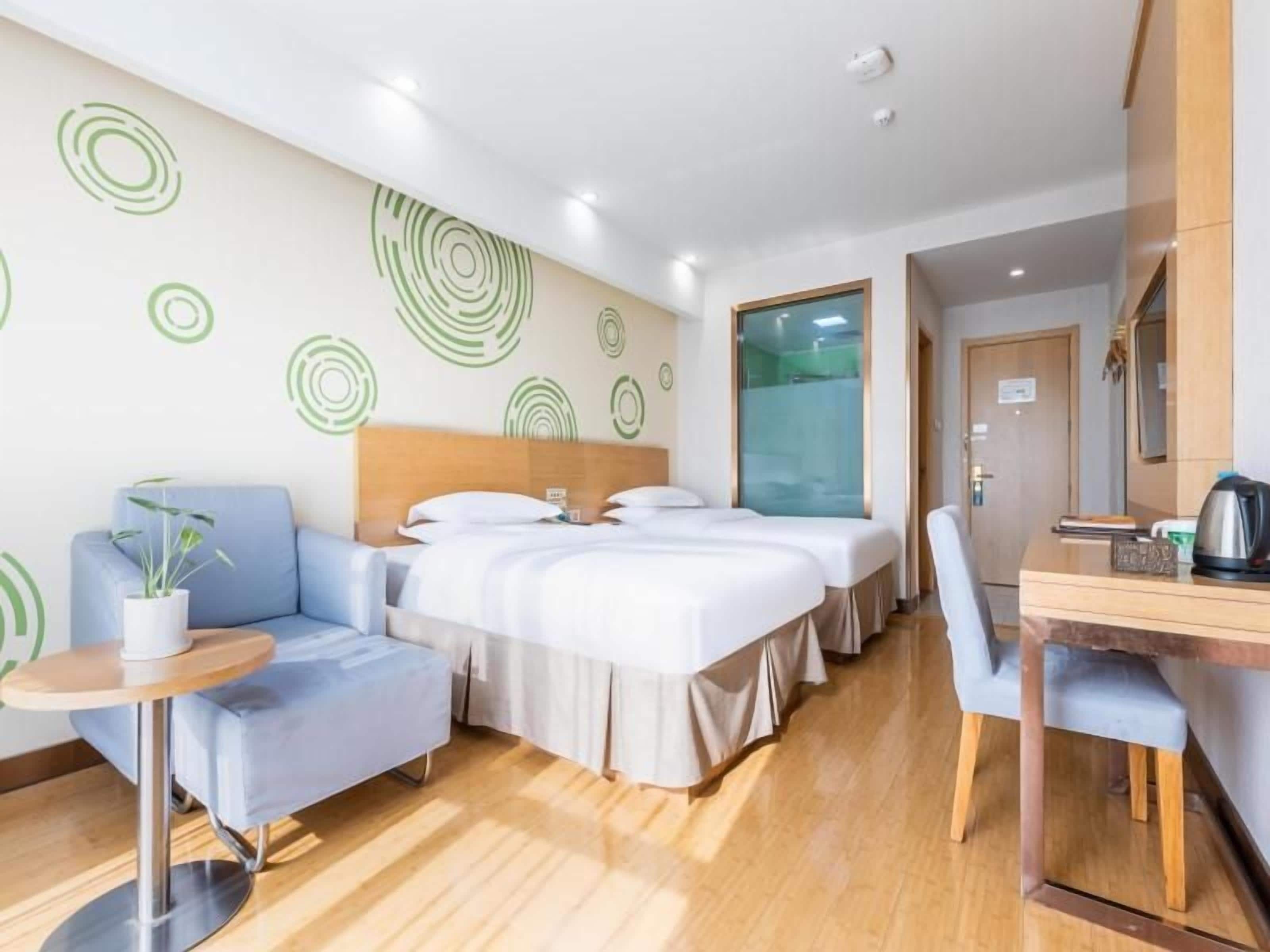 A hotel room with a bed, a desk, a chair, a small table, and a potted plant.