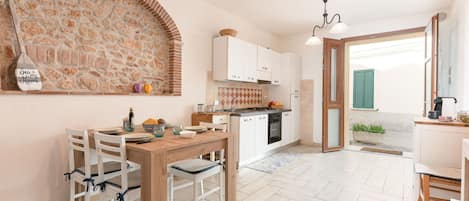 Une cuisine chaleureuse avec un mur de pierres apparentes derrière l’évier, une table à manger en bois et des armoires blanches.