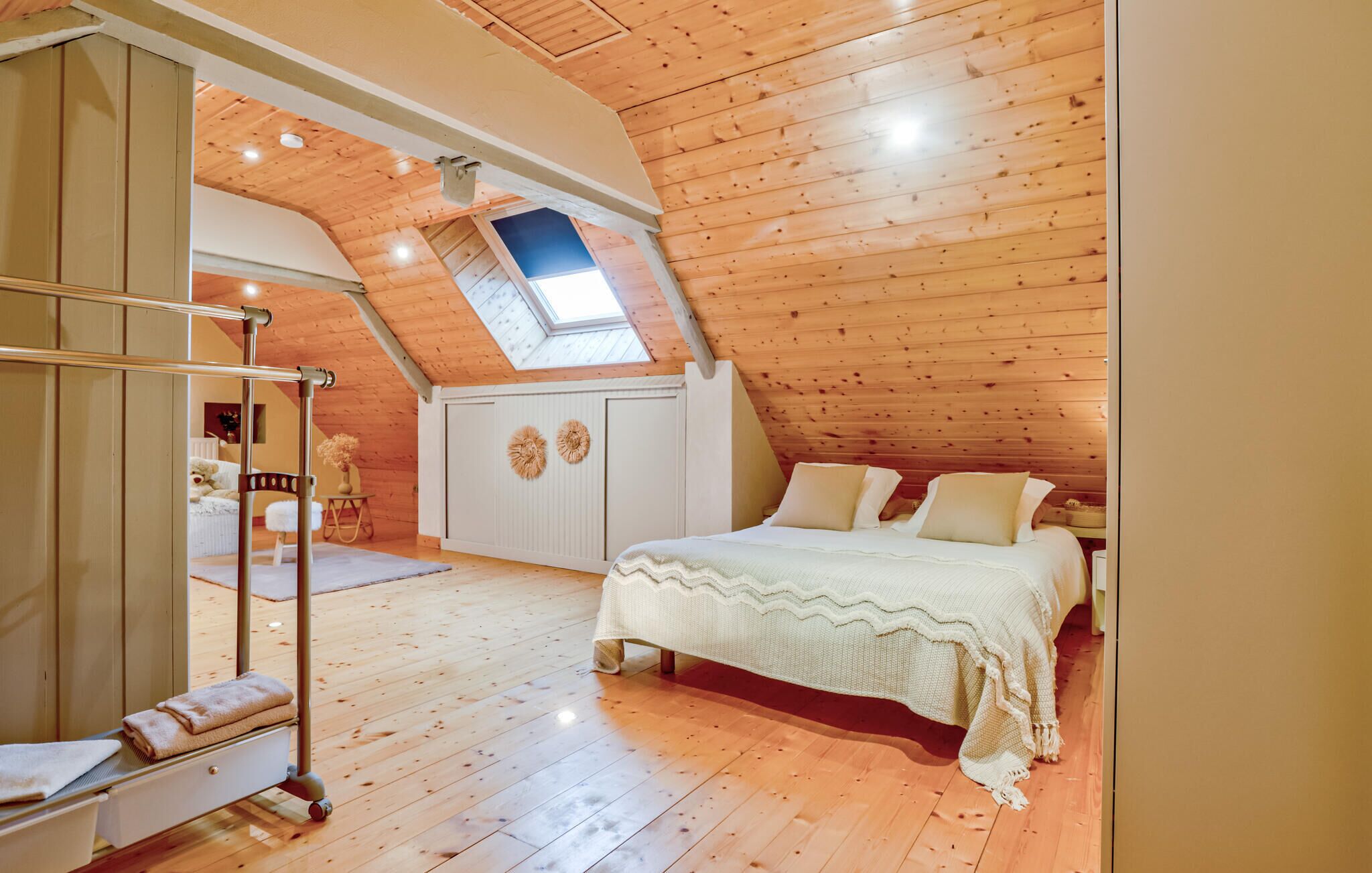 A cozy attic bedroom with a sloped ceiling, wooden walls, a skylight, a bed with a knitted blanket, and a small table with a vase.