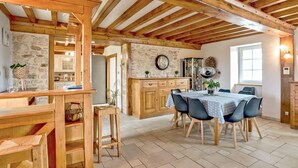 A rustic interior with exposed wooden beams, a stone wall, a wooden dining table with chairs, and a kitchen area with cabinets and a refrigerator. - Beautiful home in l'étang-bertrand (L'Étang-Bertrand)