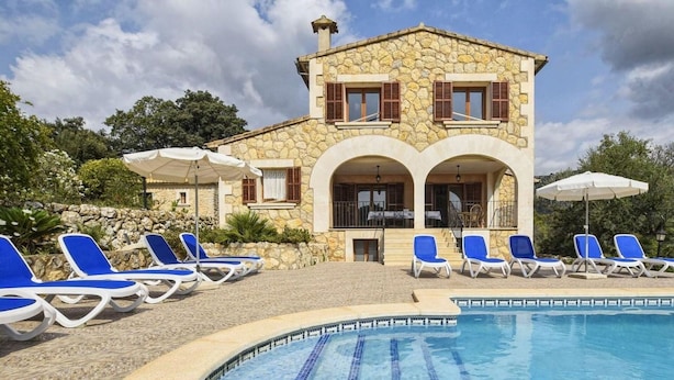 A stone building with a swimming pool, blue lounge chairs, and umbrellas.