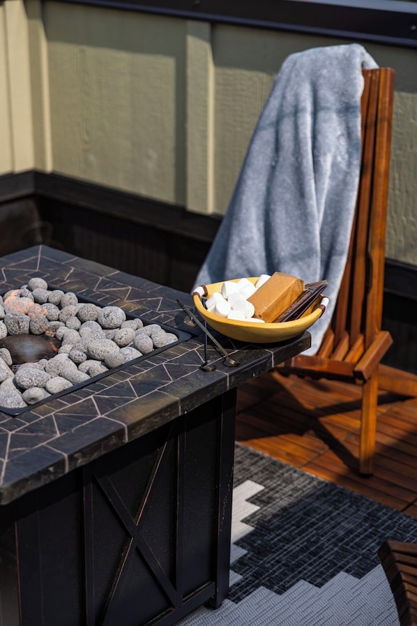 A stone fire table with a bowl of marshmallows and a box of chocolate, a wooden chair with a towel, and a patterned rug on a wooden floor.