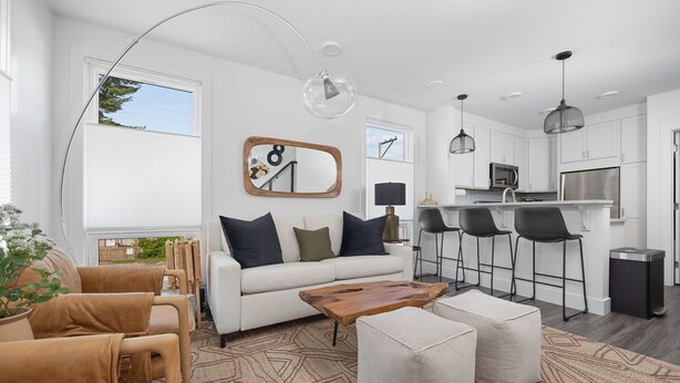 Un salon moderne avec un canapé, un pouf et une table basse, un coin repas avec des tabourets de bar, et une cuisine avec des armoires blanches et des appareils en acier inoxydable.