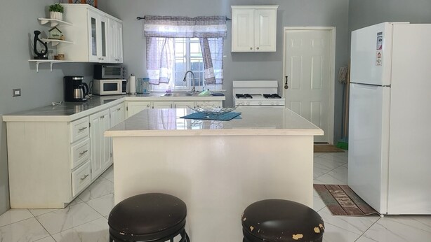 Une cuisine avec des meubles blancs, un îlot central, deux tabourets, un réfrigérateur et un luminaire suspendu.