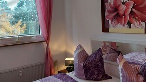 A bedroom with a large window, a radiator, a bedside table with a lamp, and a bed with multiple pillows.