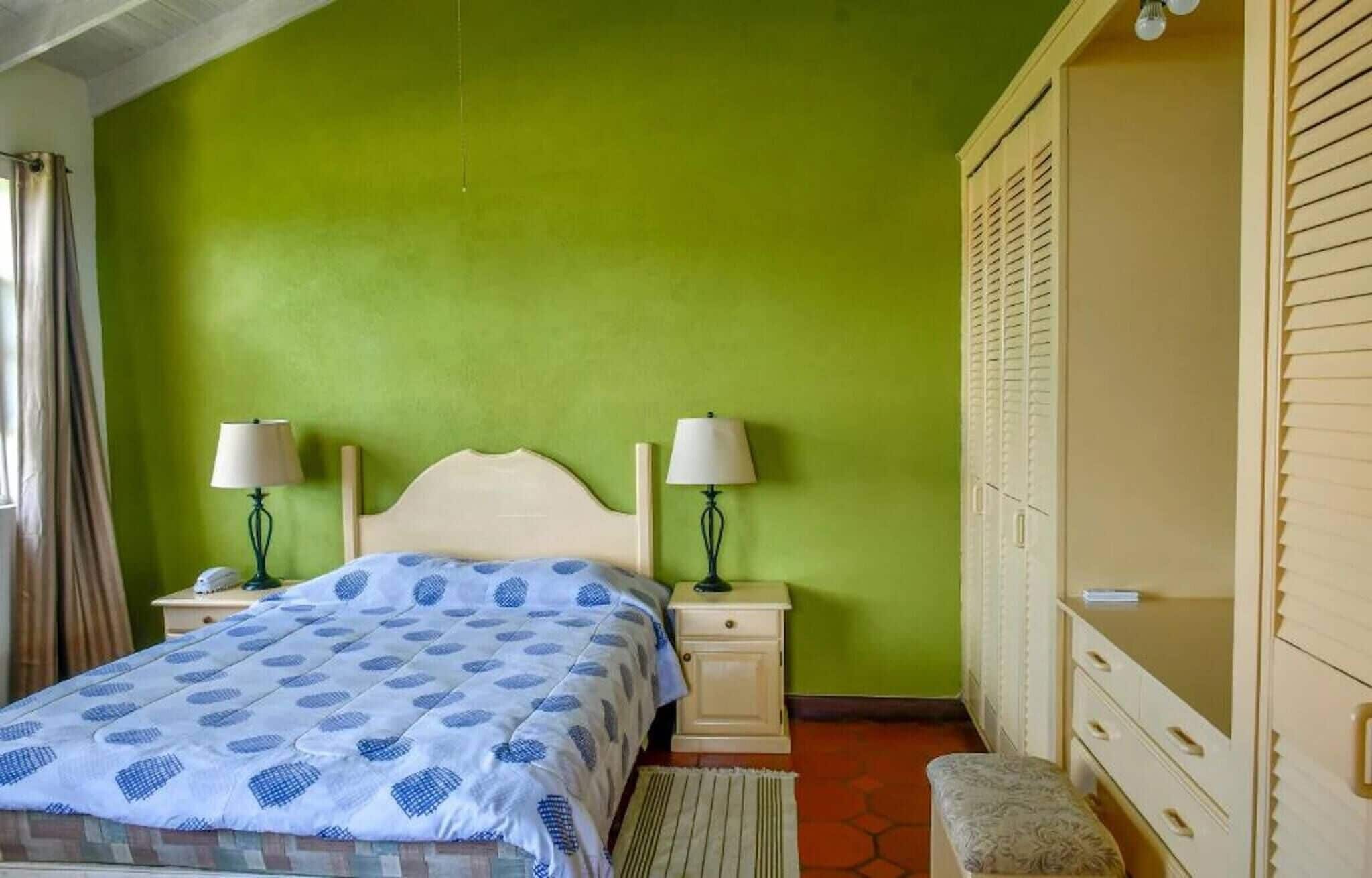 A bedroom with a bed, two bedside tables with lamps, a bench, a wardrobe, and a green wall.
