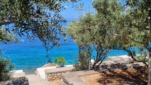 Un escalier en pierre mène à une terrasse d’où l’on peut admirer une mer d’un bleu profond, le tout encadré par des oliviers qui agrémentent le paysage.