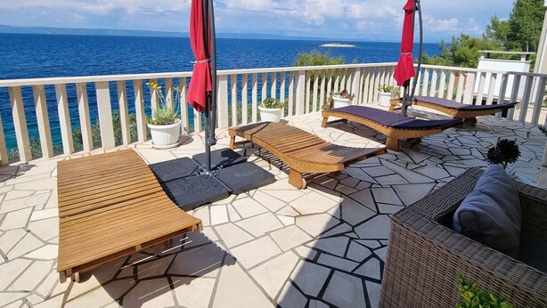 Une terrasse dotée de chaises longues en bois et de parasols rouges, offrant une vue sur une étendue d’eau et une île lointaine.