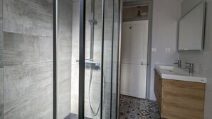 A modern bathroom with a glass-enclosed shower, a white door, a wooden vanity, and a tiled floor. - Appartement en Entier 2 Chambres 6 Couchages (Nancy)