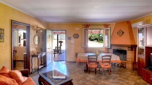 Ein gemütliches Wohnzimmer mit einer Couch, einem Couchtisch und einem Essbereich mit Stühlen und Tisch. Ein Kamin sorgt für Wärme und durch ein Fenster bietet sich ein Blick ins Freie.