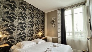 A hotel room with a large bed, a nightstand, a wall with a fern pattern, a window with curtains, and a view of buildings outside.