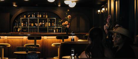 Un coin bar à la lumière tamisée, avec des meubles en bois, un comptoir où sont exposées des bouteilles et un vase décoratif rempli de fleurs.