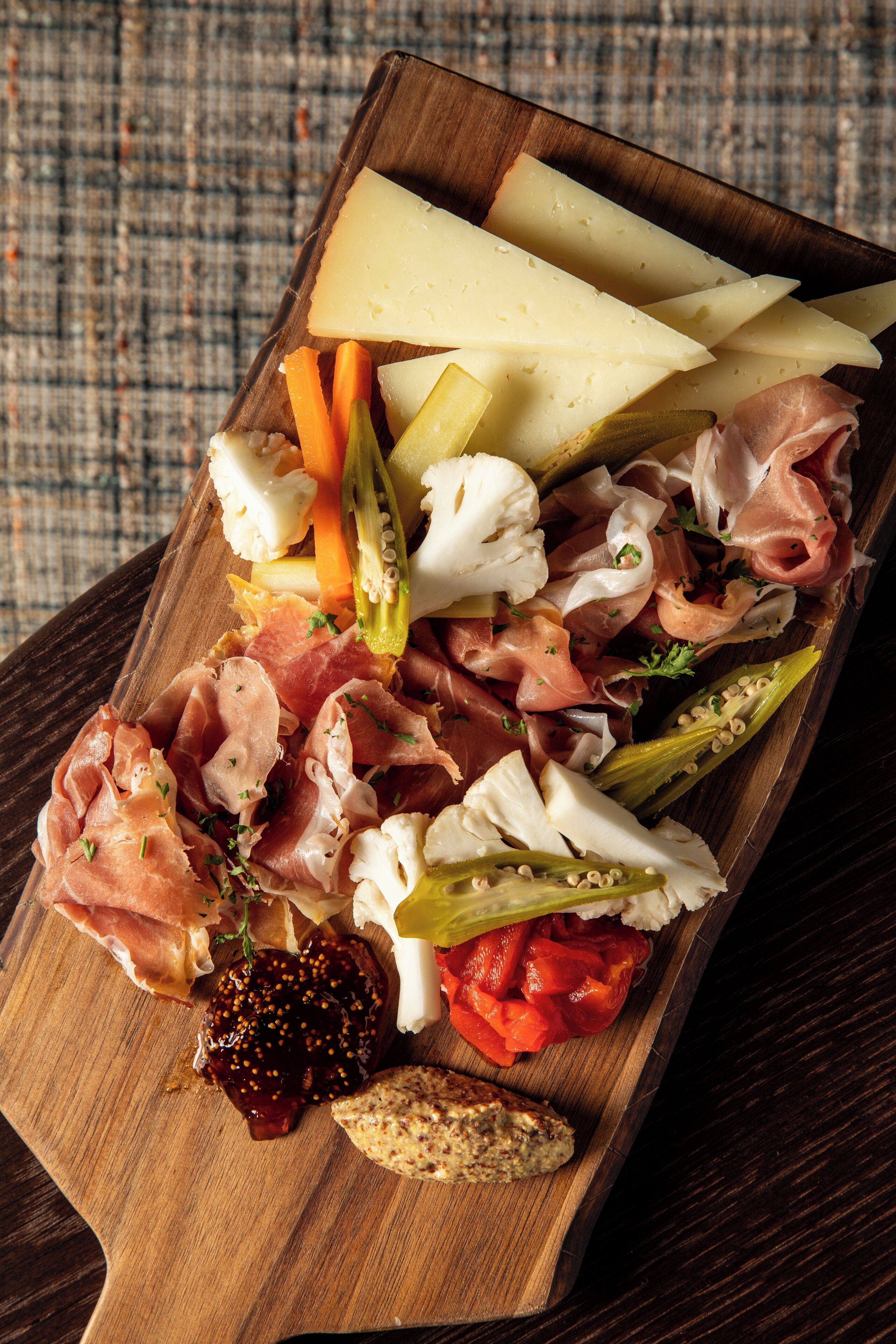 A wooden board with various cheeses, cured meats, vegetables, and bread.