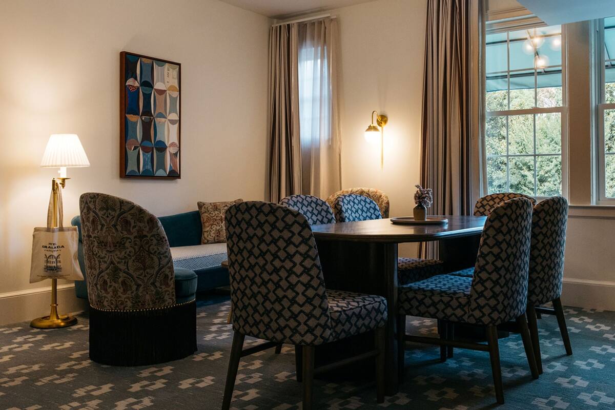 A room with a dining table surrounded by patterned chairs, a sofa with cushions, a lamp, and a painting on the wall.
