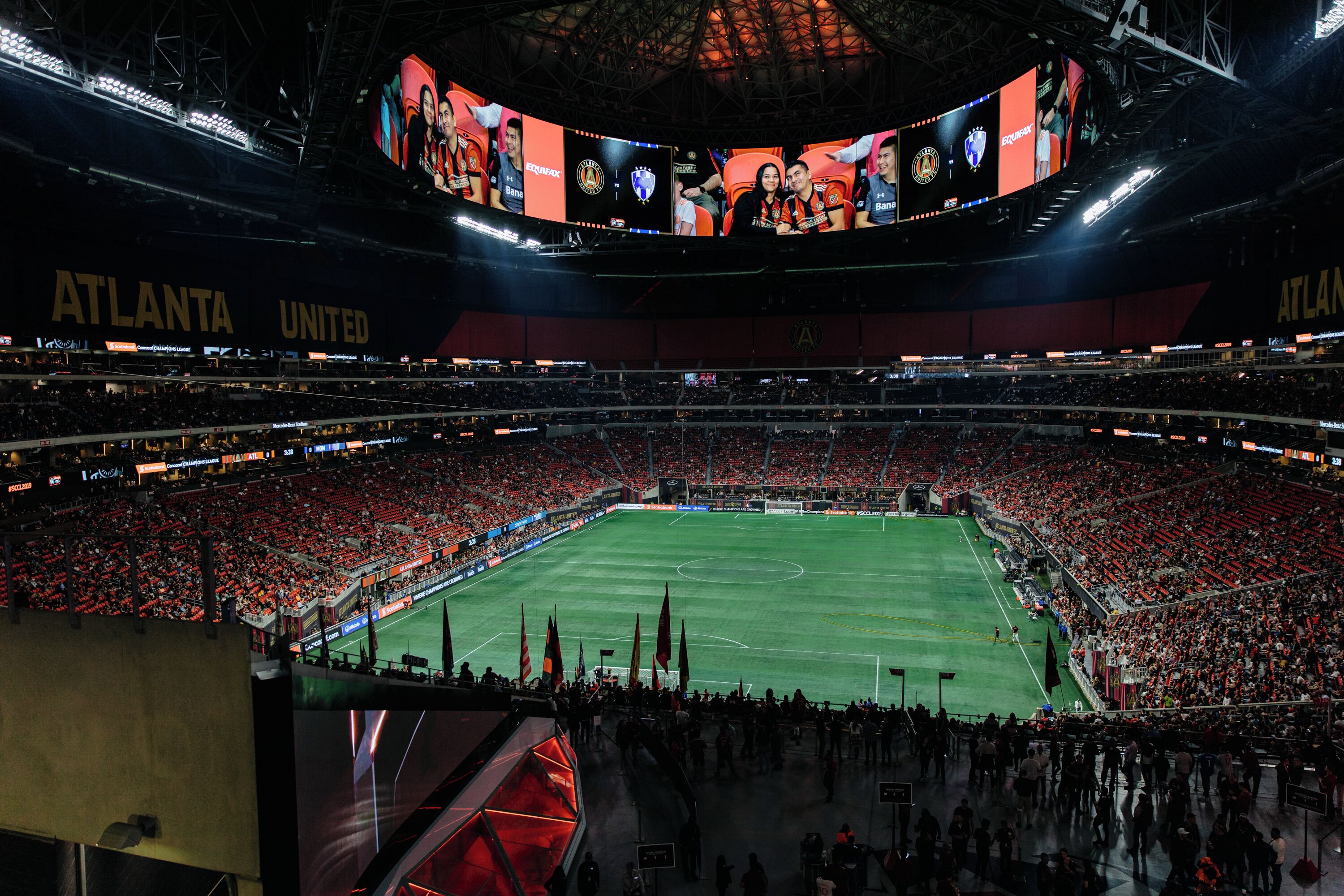 A large stadium with multiple tiers of seating, a central field, and a large digital display above the field.