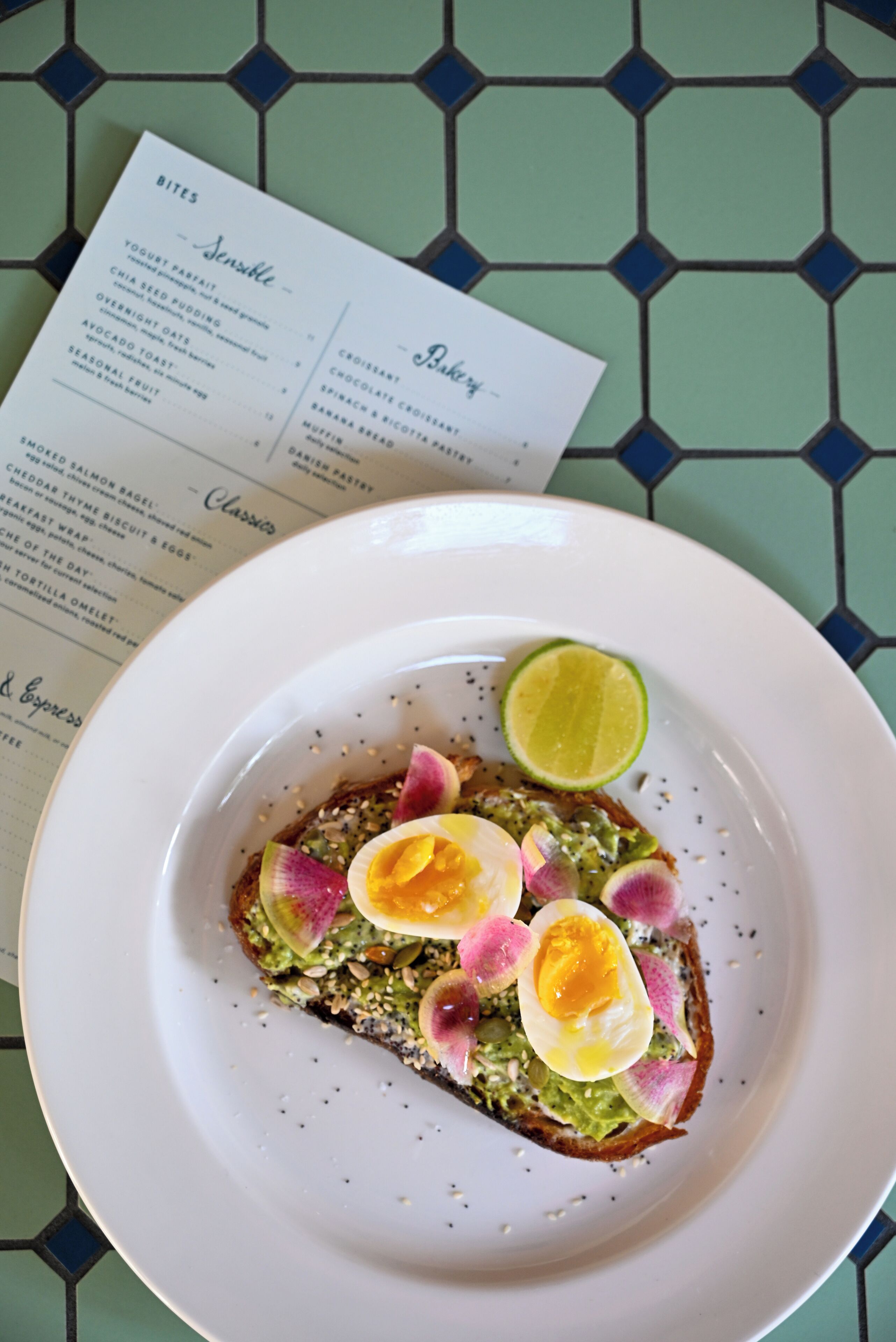 A plate with avocado toast topped with radish slices and a lime wedge, on a table with a menu.