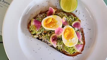 A plate with avocado toast topped with radish slices and a lime wedge, on a table with a menu.