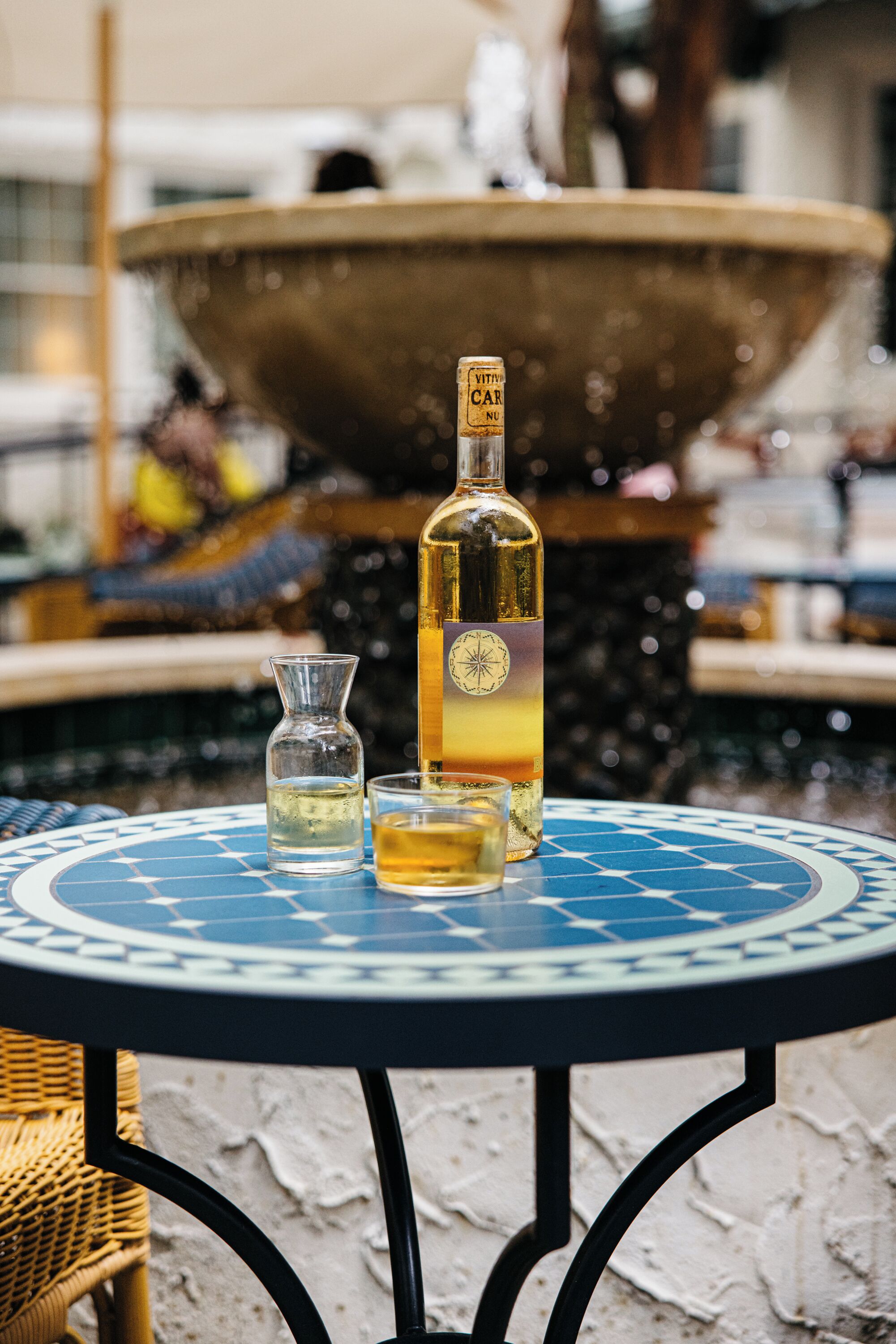 Ein Tisch mit einer Flasche und zwei Gläsern, im Hintergrund eine Springbrunnen und ein Korbstuhl.