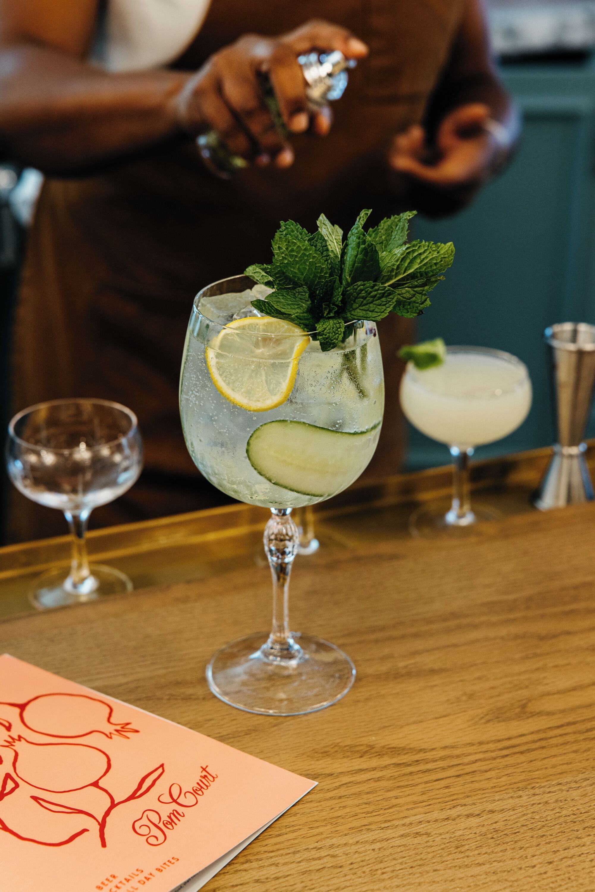 A bar setting with a drink garnished with lemon and mint, a menu, and a bartender preparing another drink.