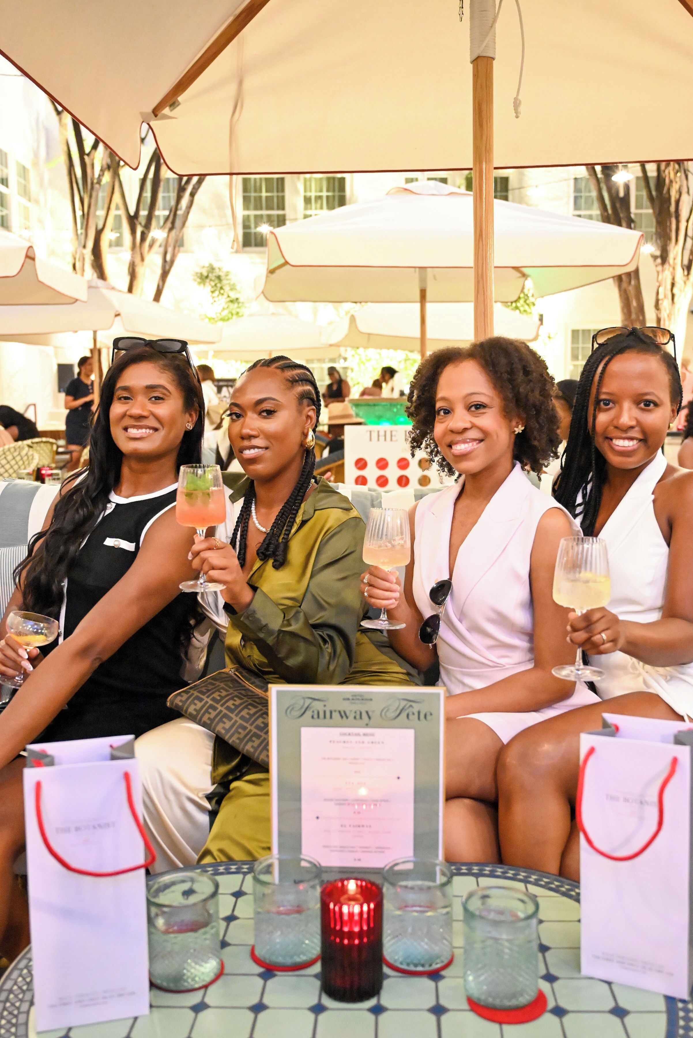 A group of people seated at an outdoor table with drinks, surrounded by umbrellas and a menu stand.