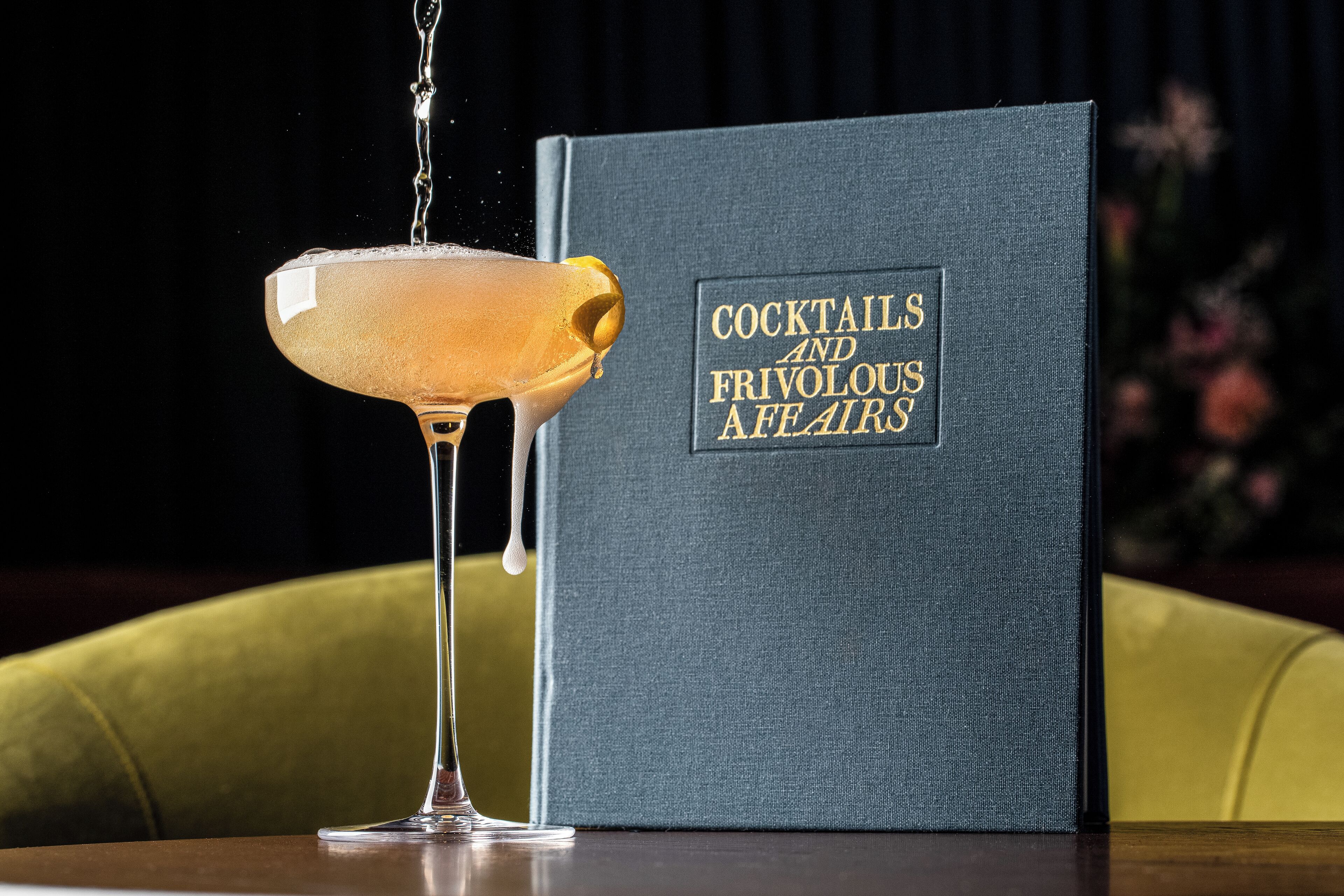 A cocktail glass with a golden drink and a book titled "COCKTAILS AND FRIVOUS AFFAIRS" beside it.