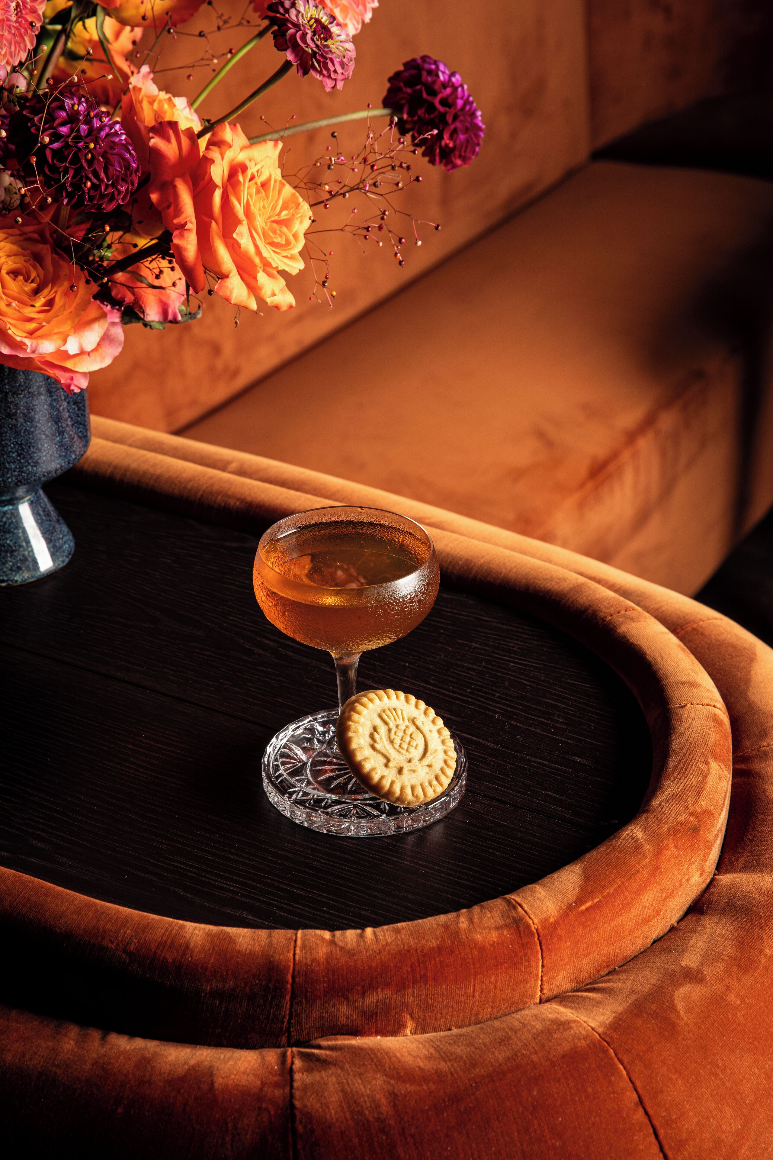 Un bicchiere da cocktail su un sottobicchiere, un biscotto e un vaso con fiori su un tavolo.