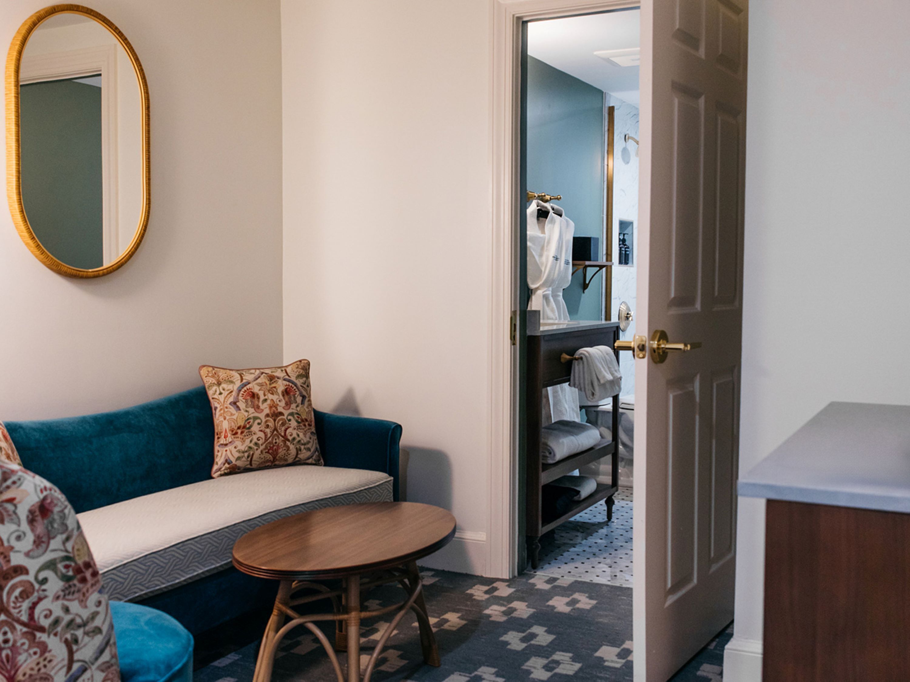 A hotel room with a sofa, a small round table, a mirror, and a door leading to a bathroom with a towel rack.