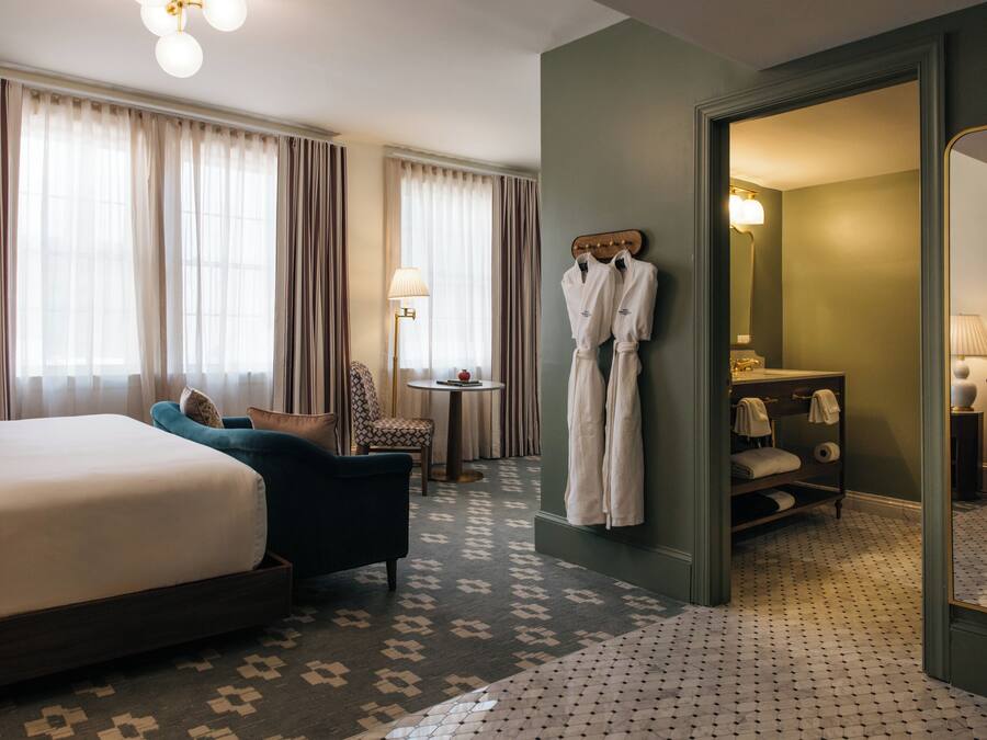 A hotel room with a large bed, a sitting area with a chair and a small table, a bathroom with a mirror and towel rack, and a patterned floor.