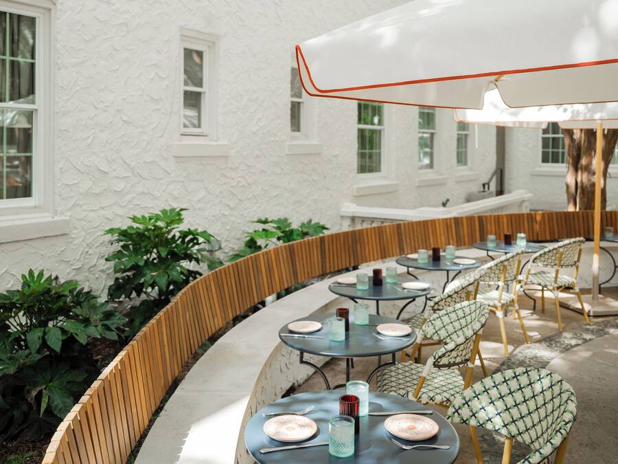 Un patio al aire libre con mesas y sillas redondas, rodeado por un edificio blanco con ventanas.