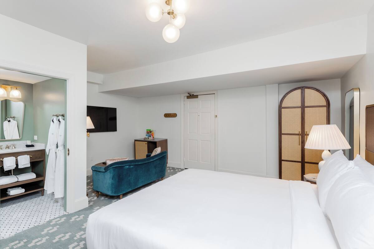A hotel room with a large bed, a vanity with a mirror, a blue armchair, a wooden wardrobe, and a door with a window.