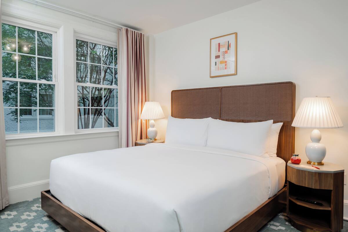 A neatly made bed with white linens, two bedside lamps, a framed picture on the wall, and a window with curtains.