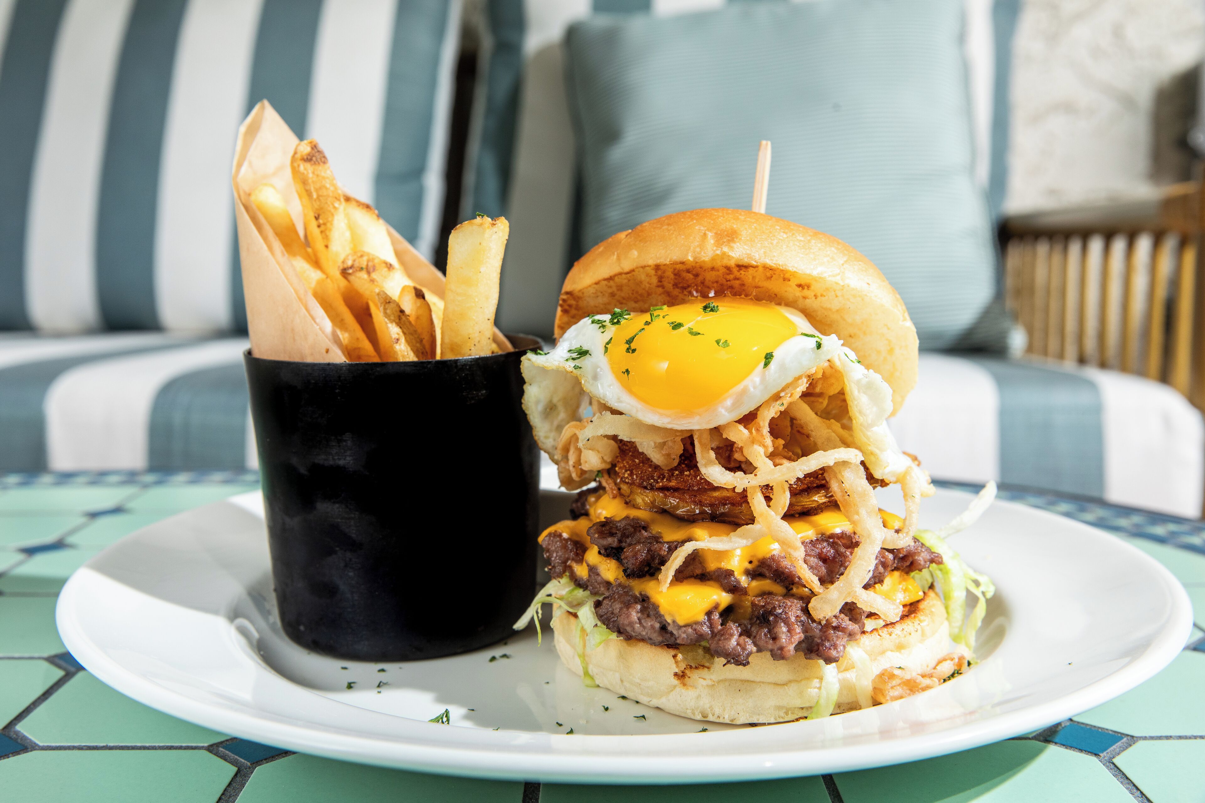 Une assiette avec un hamburger et des frites, une tasse noire et, à l’arrière-plan, un canapé à rayures bleues et blanches.
