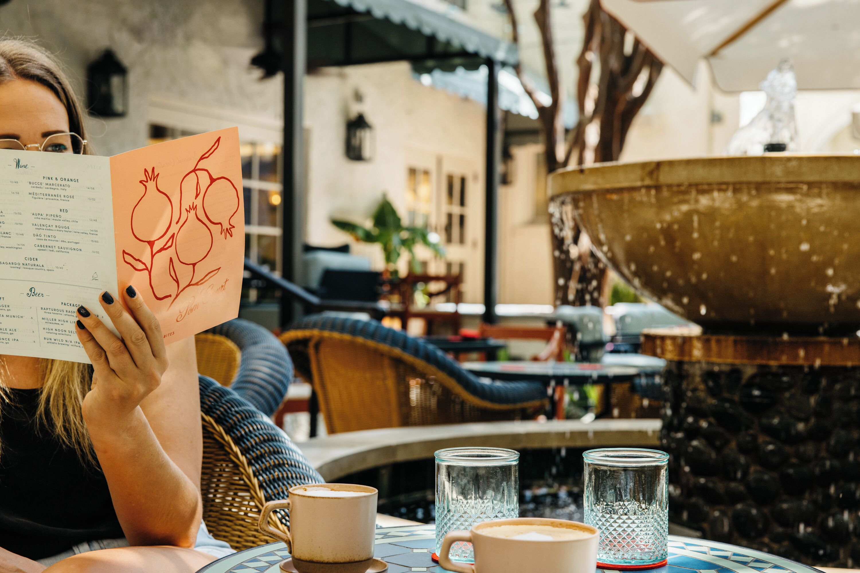 Una persona leyendo un menú en un café al aire libre con una fuente y mobiliario de mimbre.