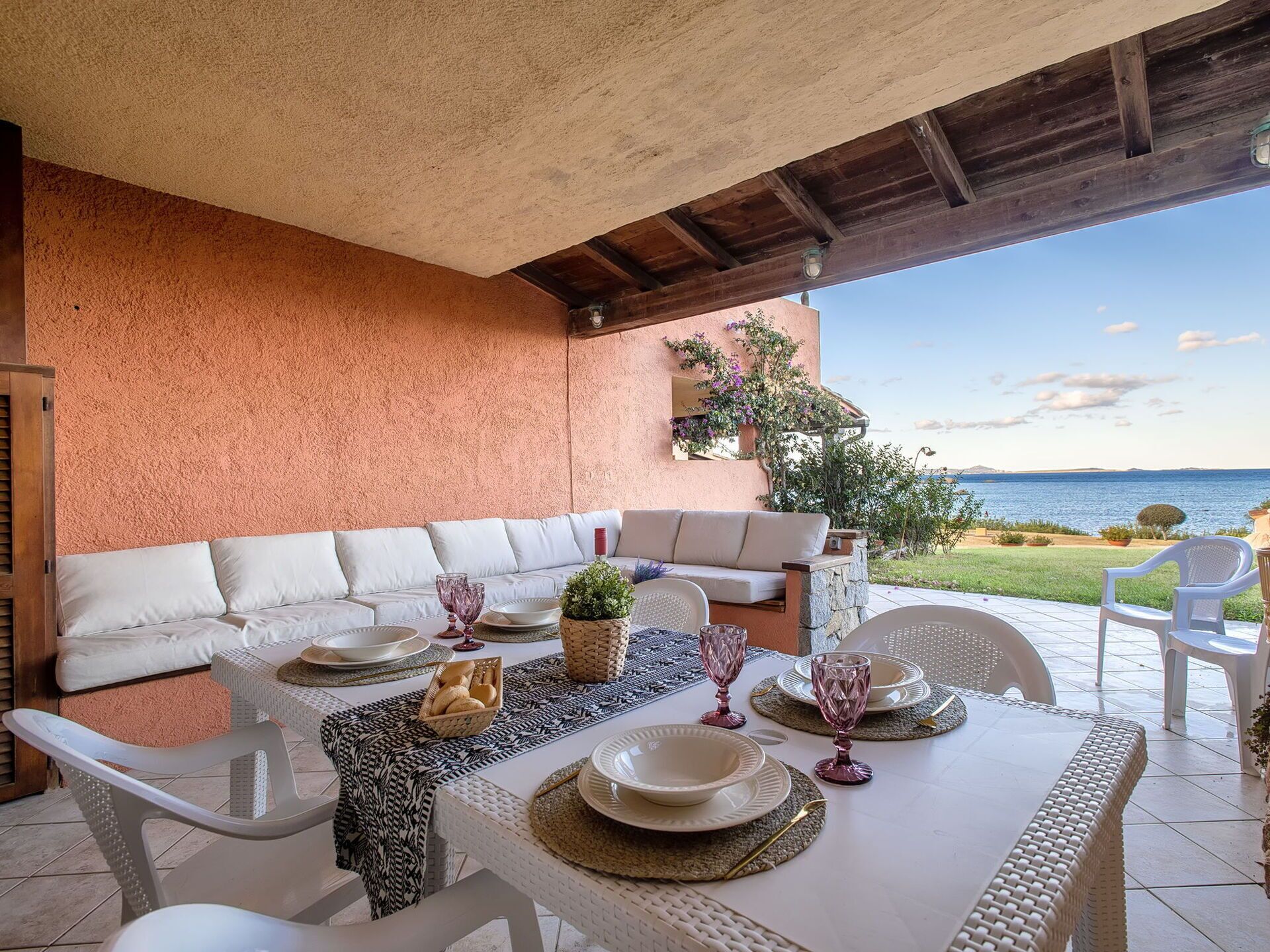 An outdoor dining area with a white table set for four, a white sofa, and a view of the sea.