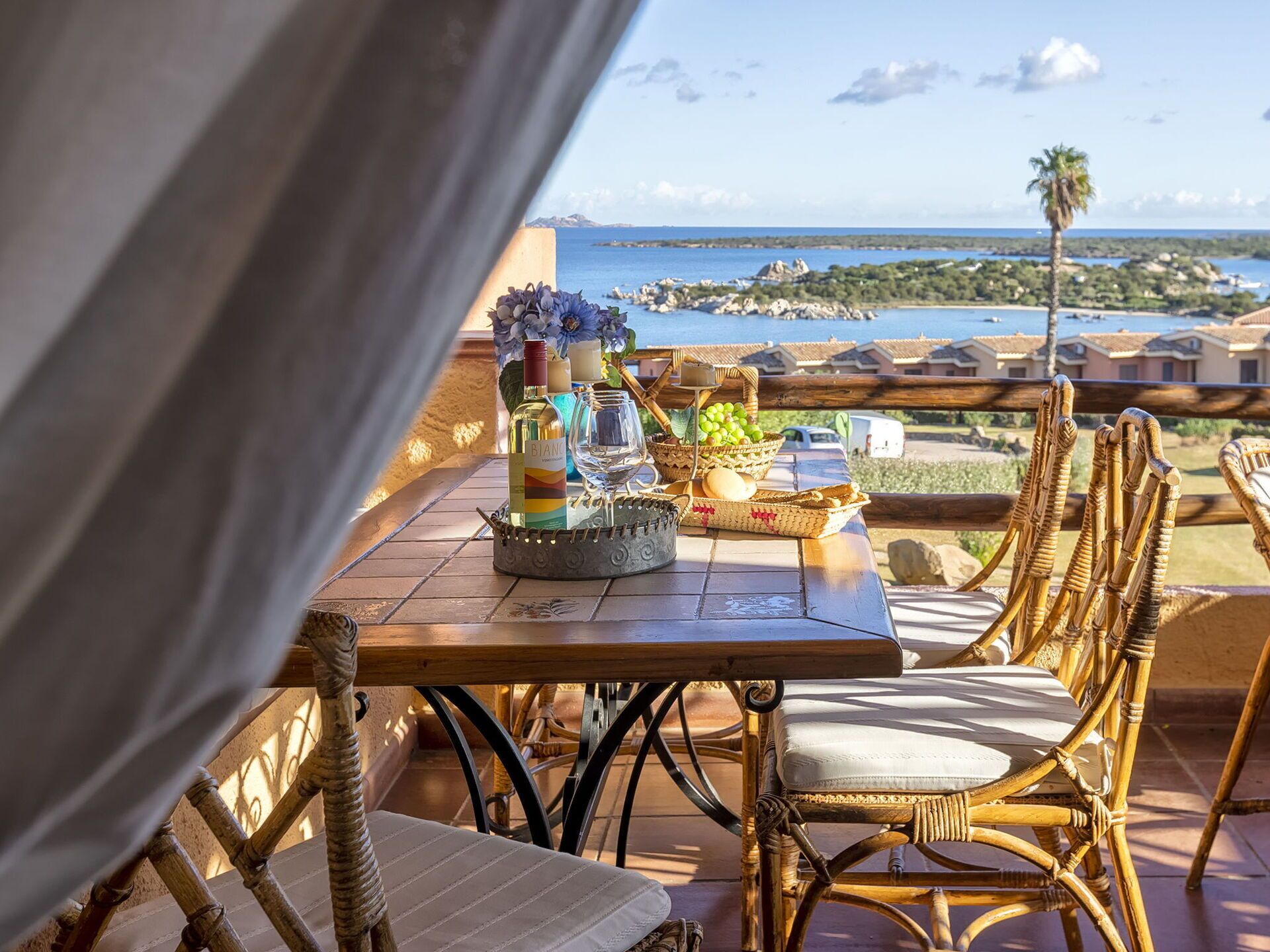 Een tafel gedekt met een fles wijn, glazen en een mand met druiven op een terras met uitzicht op de kust.