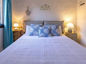 Een net opgemaakt bed met witte en blauwe kussens, twee nachtlampjes en een decoratie aan de muur.