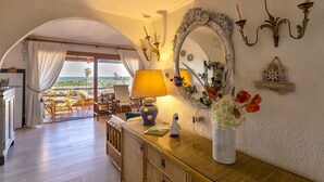 A room with a wooden floor, a sofa, a table with a lamp, a mirror, and a view of the outdoors. - Lovely apartment for 5 people with A/C, pool, TV, terrace and pets allowed (Olbia)