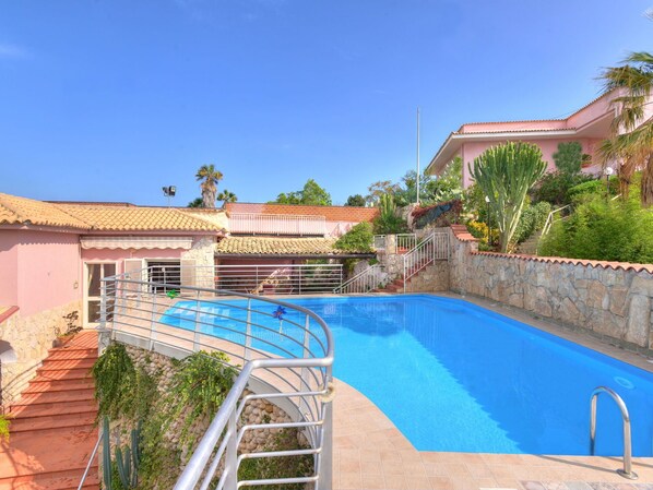 A swimming pool with a metal railing, surrounded by a stone wall and a pink building with a tiled roof.