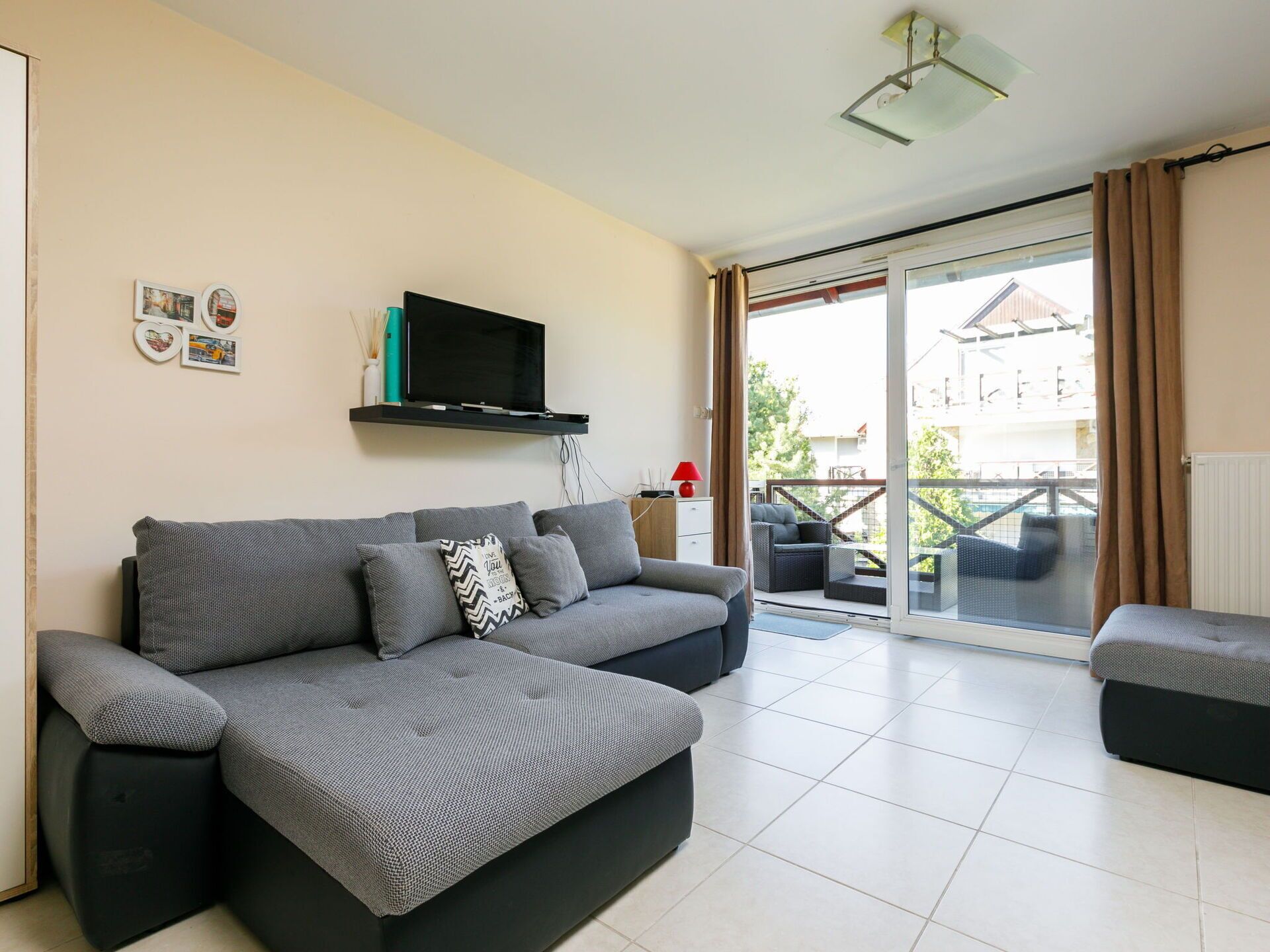 Un salon moderne avec un canapé gris, un téléviseur à écran plat fixé au mur et une porte-fenêtre coulissante donnant sur un balcon extérieur.