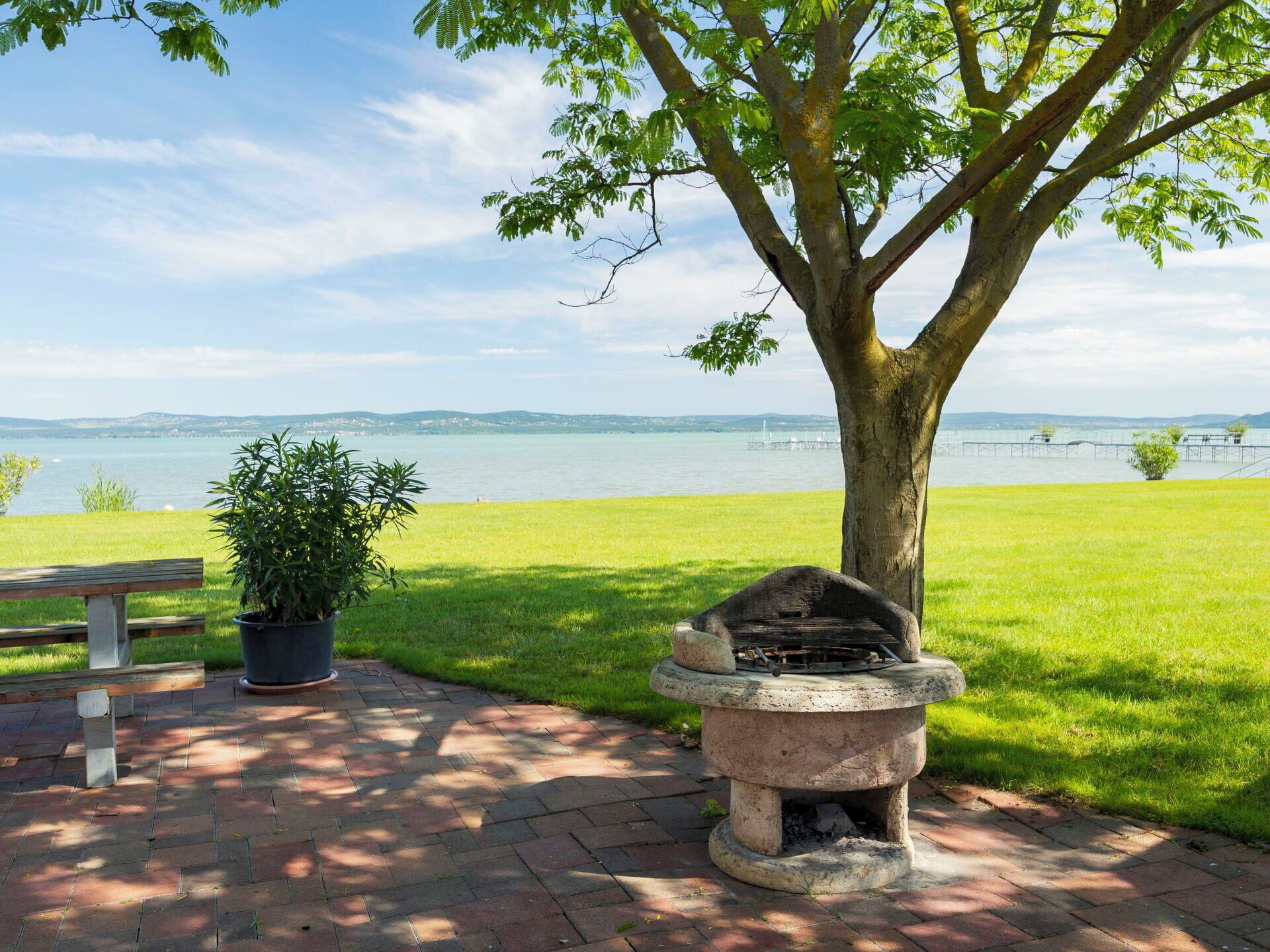 A park with a bench, a potted plant, a stone grill, and a view of a body of water.