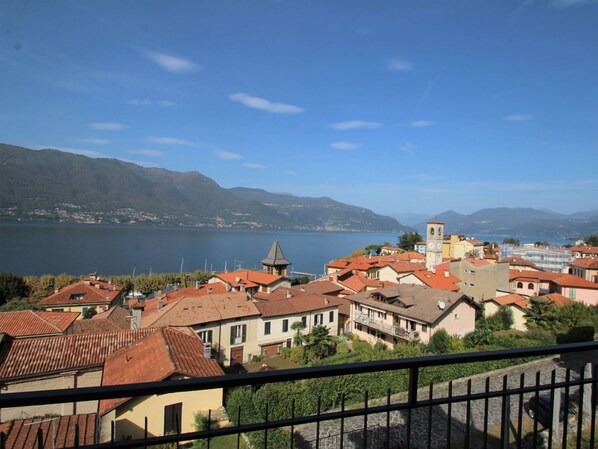 A coastal town with terracotta-roofed buildings, a prominent clock tower, and a view of the lake and mountains in the background.