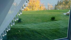 Uitzicht door een raam met een witte lijst en decoratieve details, op een grasveld met een boom in herfstkleuren, een waterpartij en heuvels in de verte.