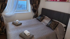 Een netjes opgemaakt bed met een gewatteerde spreideken, twee kussens en een opgerolde handdoek. Een schilderij van een bloem hangt aan de muur.