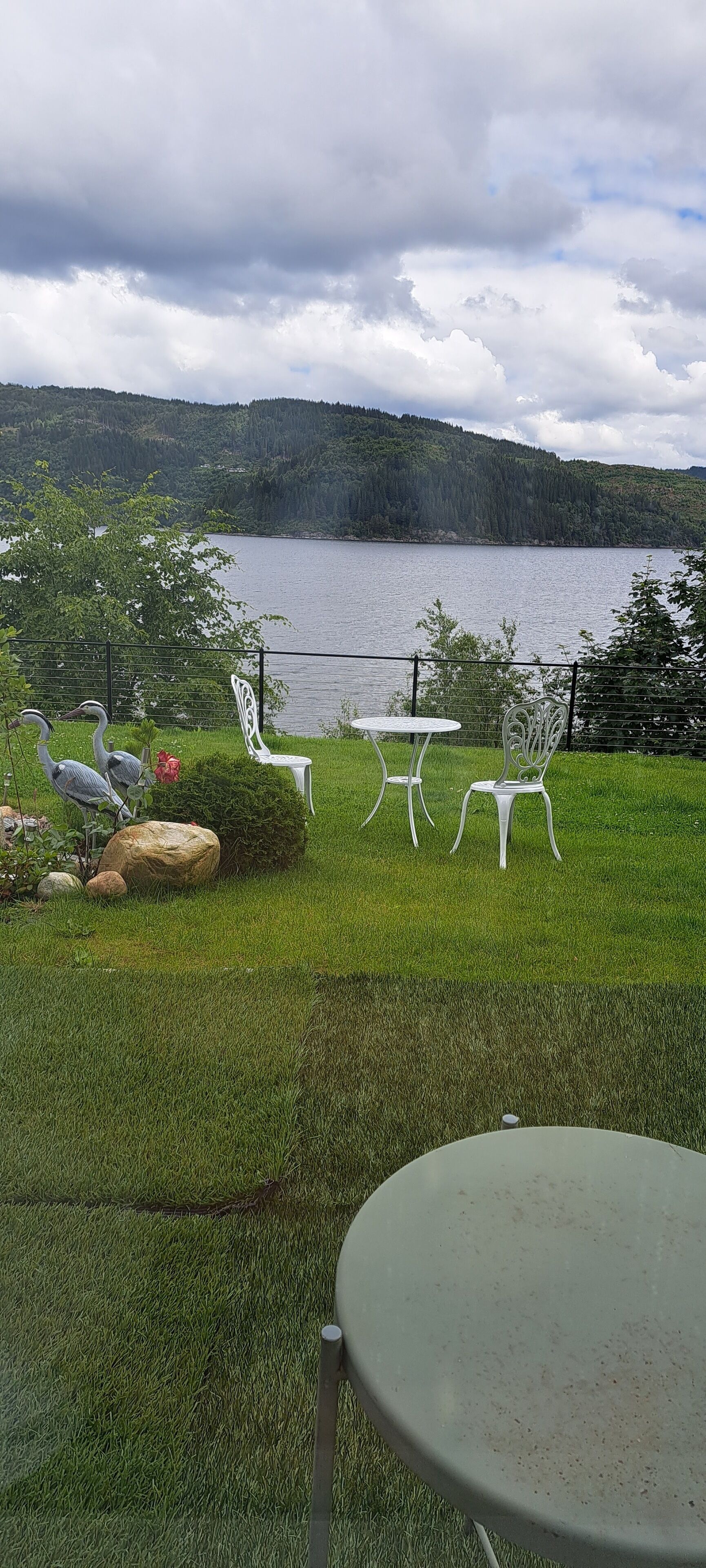 Un espacio al aire libre con césped, sillas y una mesa de metal blanco, con vistas a un lago y colinas boscosas.