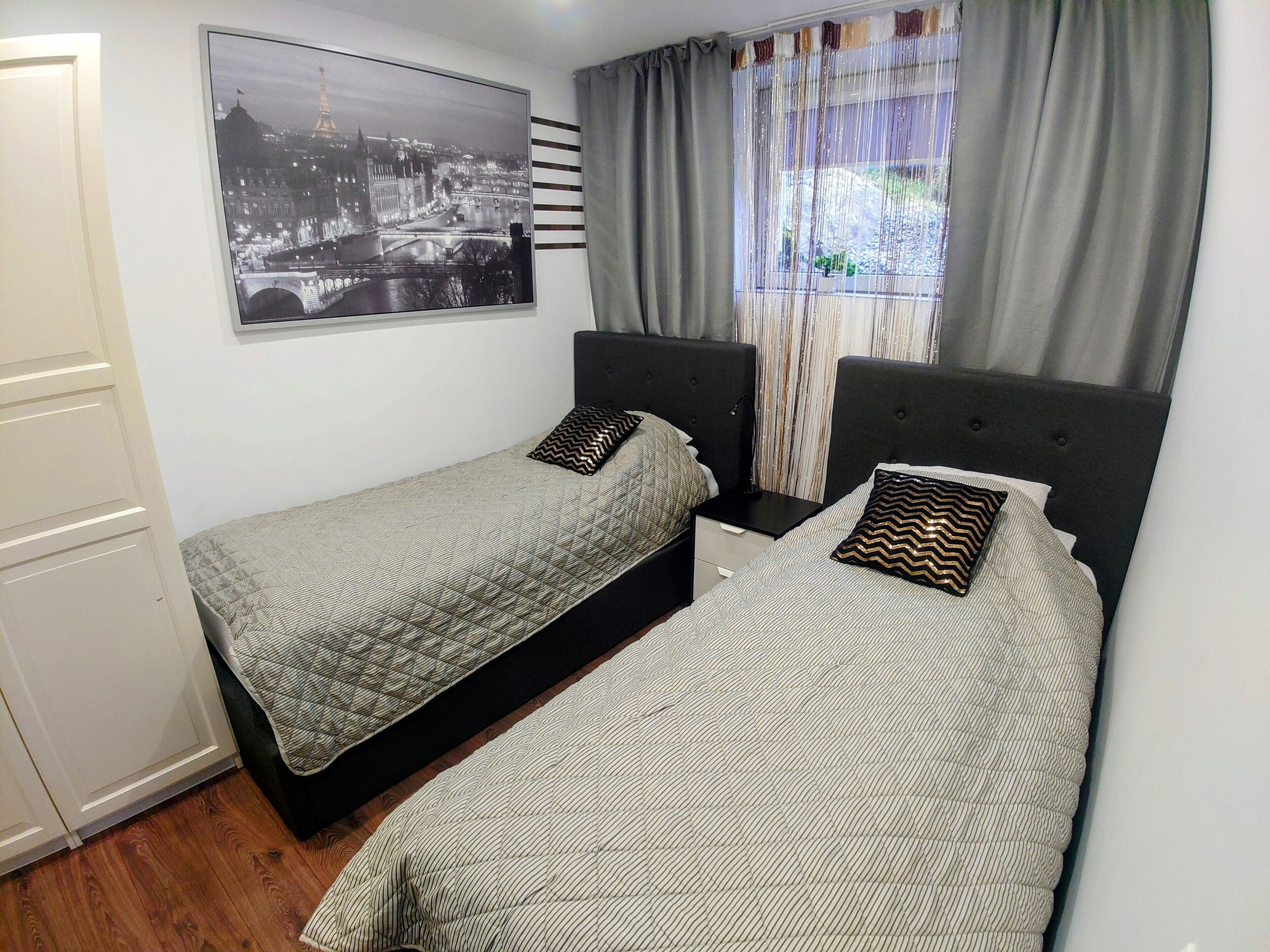 Une chambre avec deux lits, une table de chevet, une commode, une fenêtre avec des rideaux et une affiche représentant un paysage urbain accrochée au mur.