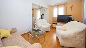 Un salon comprenant un canapé, deux fauteuils, une table basse en verre, un téléviseur et un crucifix au mur.