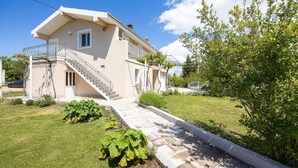 Une maison à deux étages avec un escalier menant à l’entrée, un jardin avec de la pelouse et des arbustes, et un arbre sur la droite.