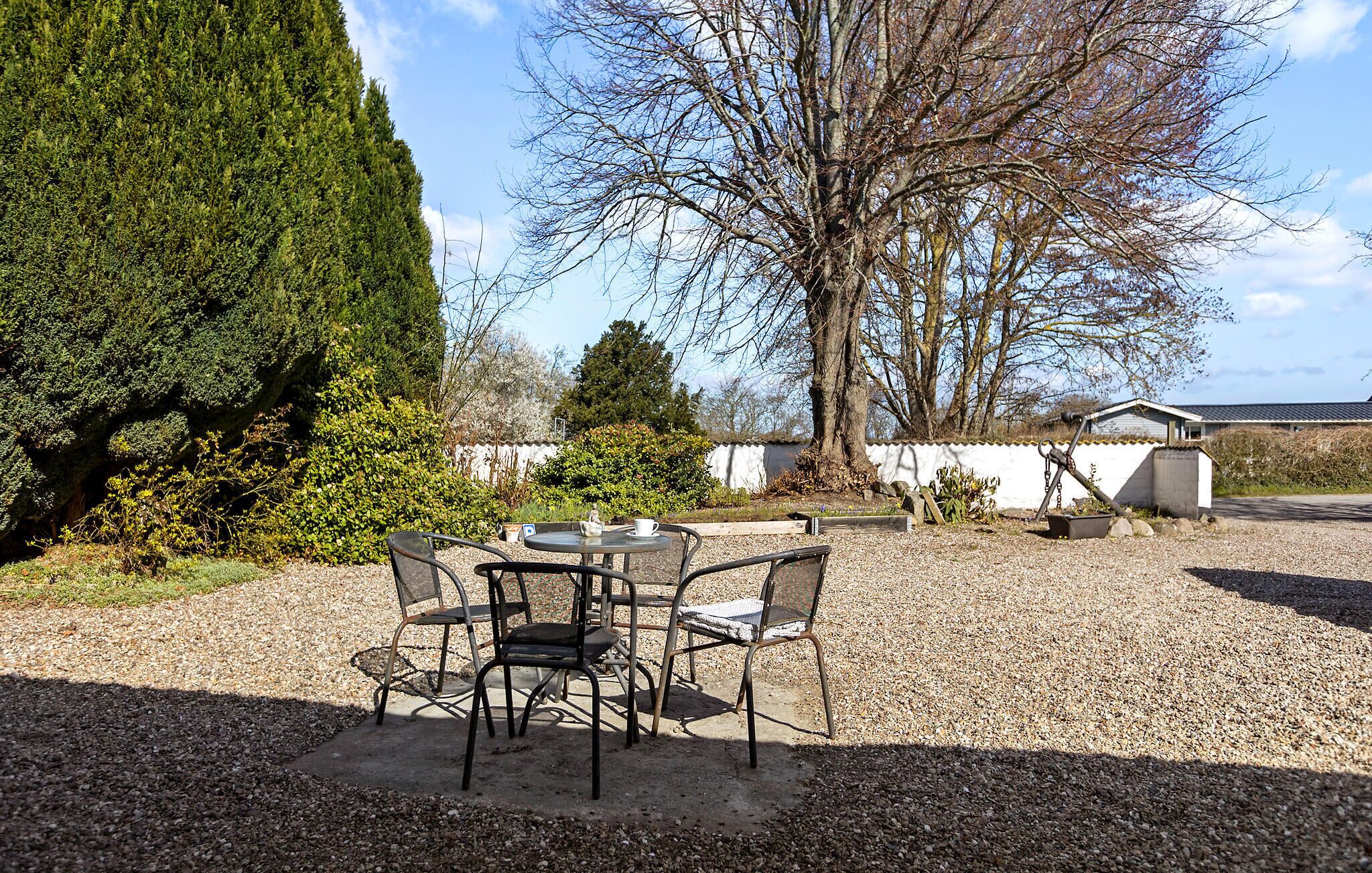 A gravel patio with a table and chairs, surrounded by trees and a white building.