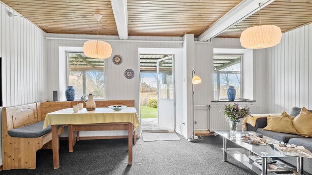 A cozy room with a wooden bench, a dining table, and a sofa. There are two windows with curtains, a door, and a small table with a vase. - 2 bedroom nice apartment in Sydals (Sydals)